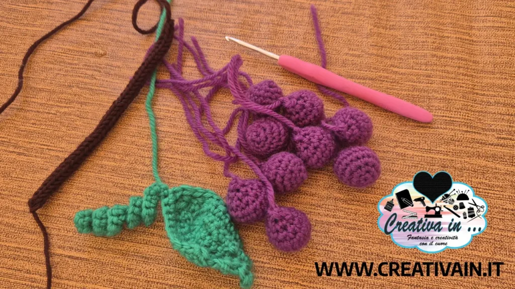 Lavori in corso per un progetto di uva amigurumi con acini viola sparsi, uncinetto rosa, foglia verde e gambo marrone su fondo in legno.
