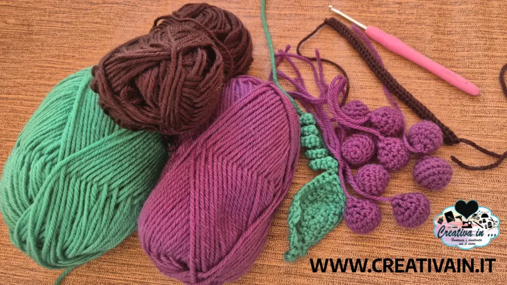 Gomitoli di lana nei colori viola, verde e marrone affiancati ai pezzi di un grappolo d'uva amigurumi in lavorazione.