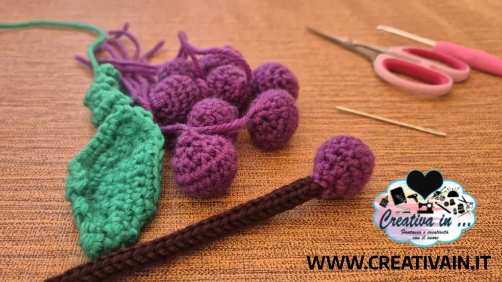 Vista ravvicinata dei singoli elementi di un grappolo d'uva amigurumi completi di acini, foglia, stelo, forbici e ago da lana.