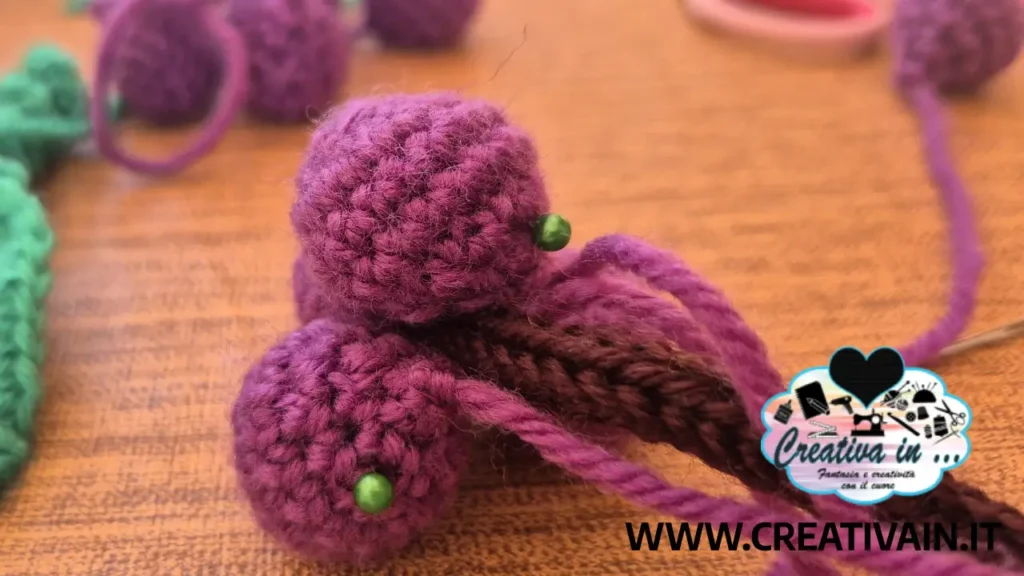 Dettaglio di acini di uva amigurumi viola fissati con spilli da testa verdi durante la fase di cucitura.