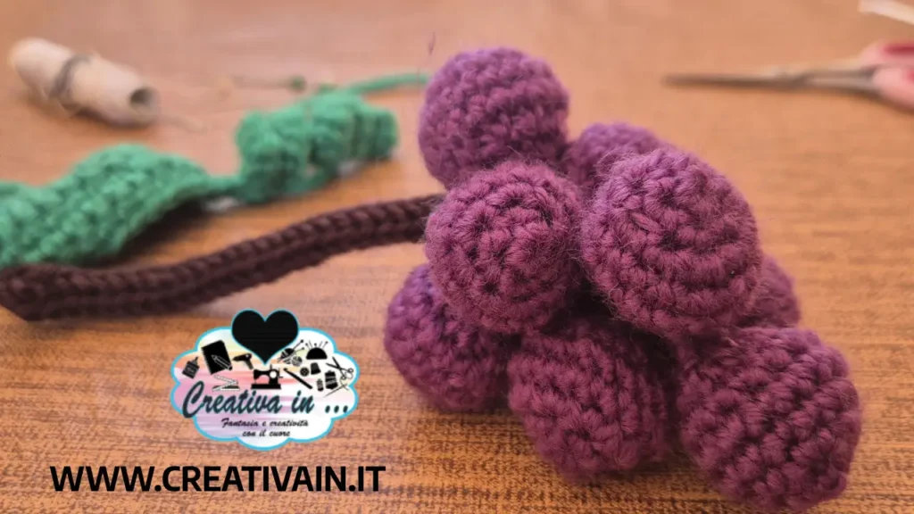 Grappolo d'uva amigurumi quasi completato con acini viola tridimensionali, foglia verde e gambo marrone